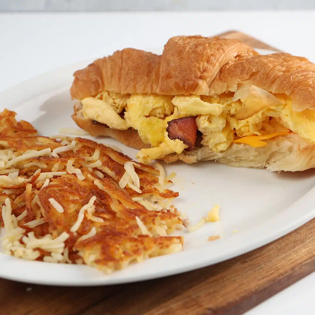 Croissant sandwich with scrambled eggs, bacon, and cheese, paired with golden-brown hash browns.