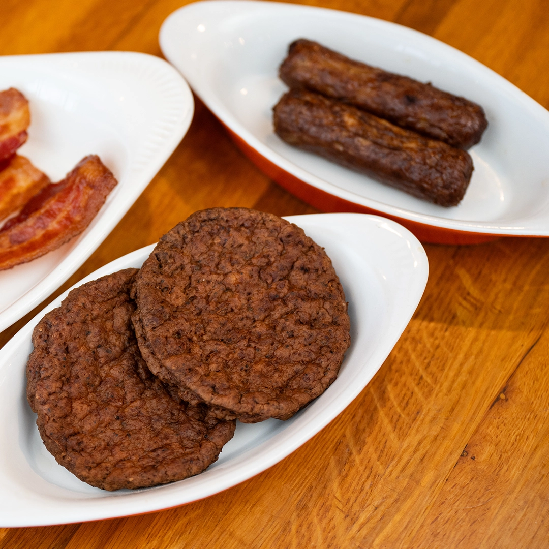 White bowls on a wooden table contain assorted breakfast meats: crispy bacon, two sausage links, and two sausage patties, all cooked to a rich brown.