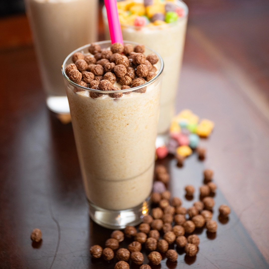 A creamy smoothie topped with chocolate cereal balls and a pink straw sits in a glass. Two more milkshakes with colorful cereal are in the background.