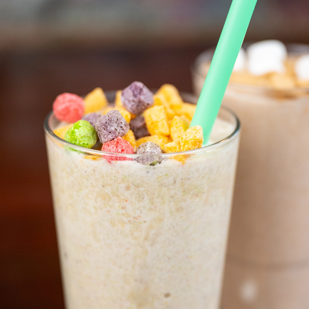 A creamy smoothie topped with colorful cereal pieces and a green straw, giving a playful and indulgent vibe. Another milkshake is blurred in the background.