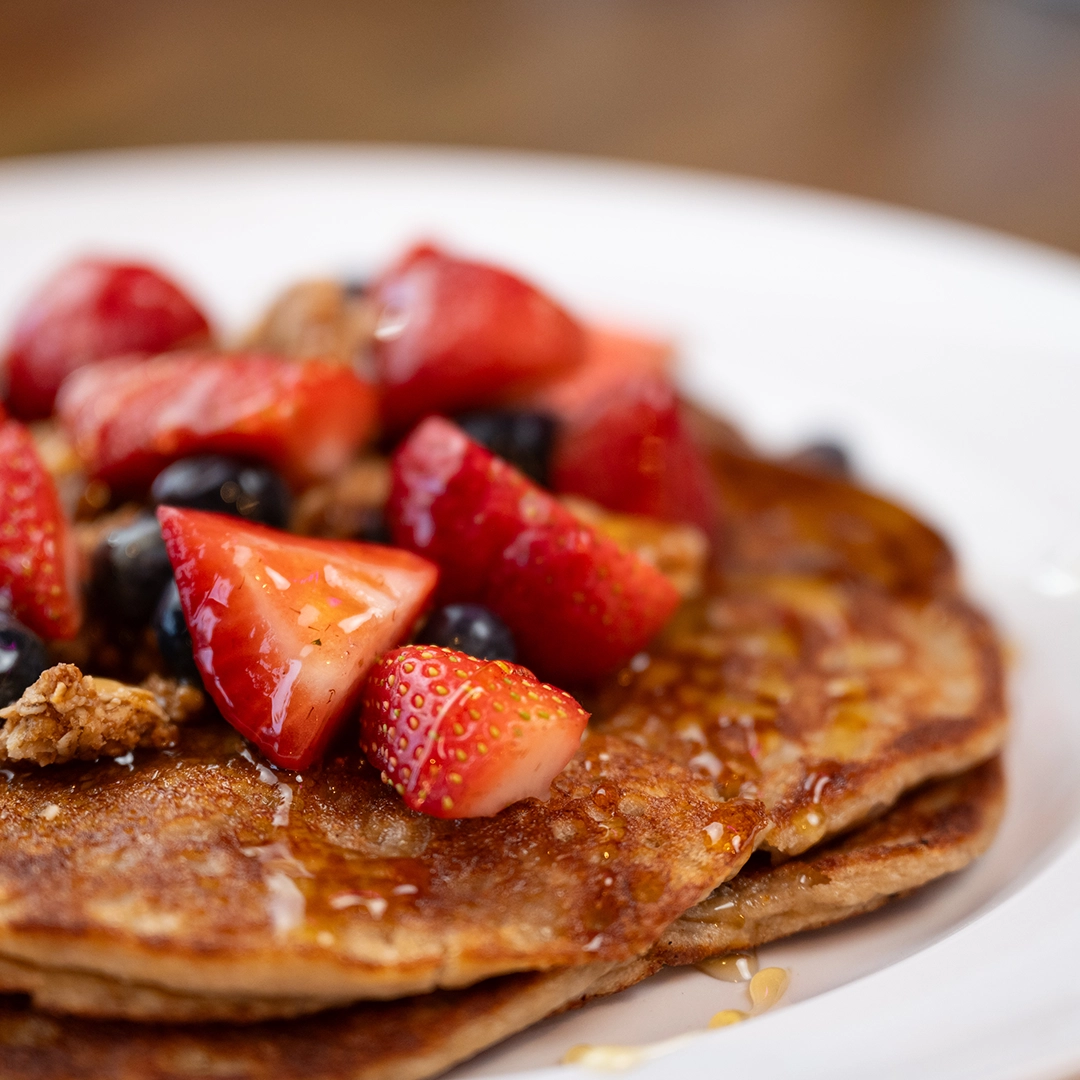 Close-up of a stack of pancakes topped with glazed strawberries, blueberries, and crunchy granola. The scene conveys a fresh, appetizing breakfast.
