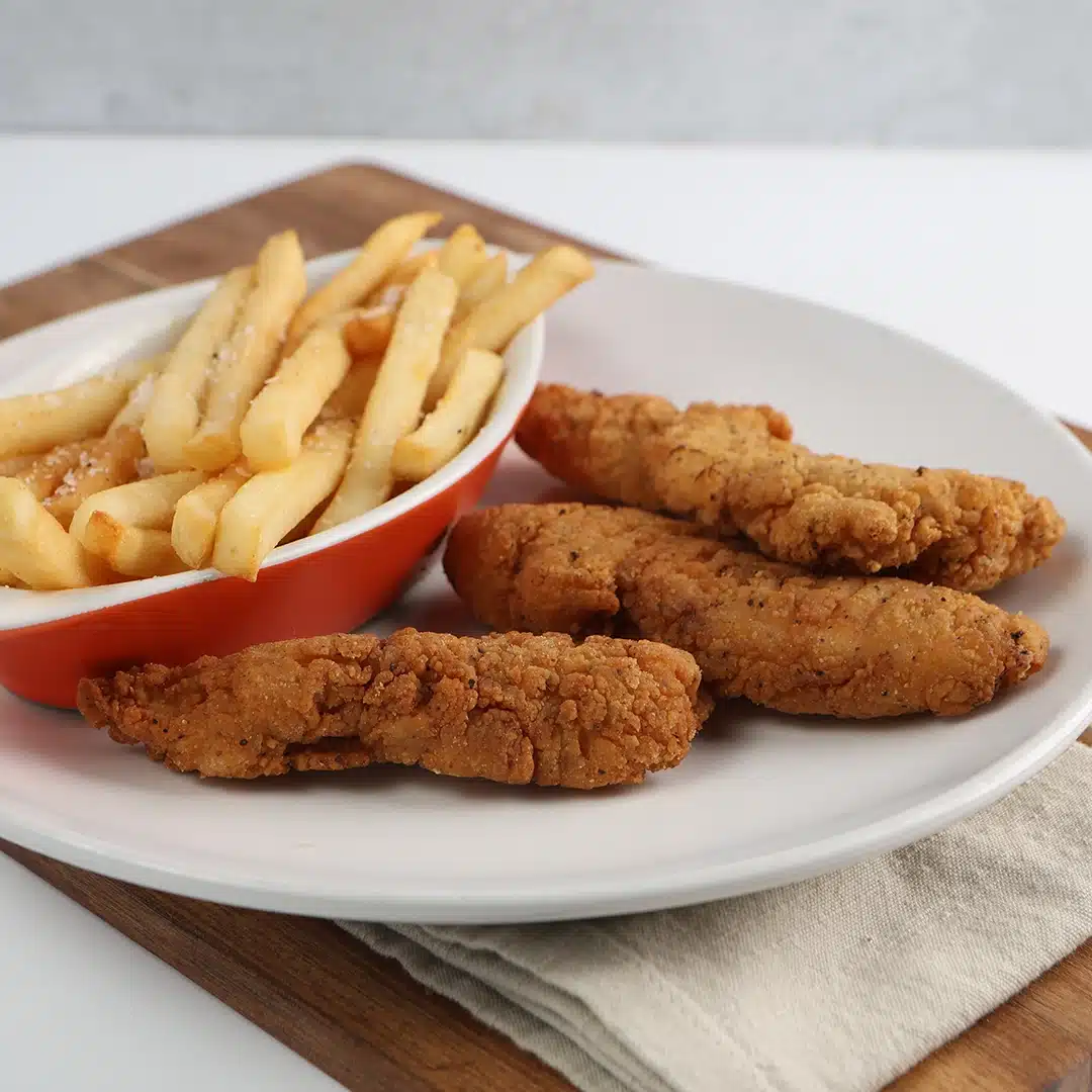 Crispy fried chicken tenders with golden French fries in a small red dish, on a wooden board.
