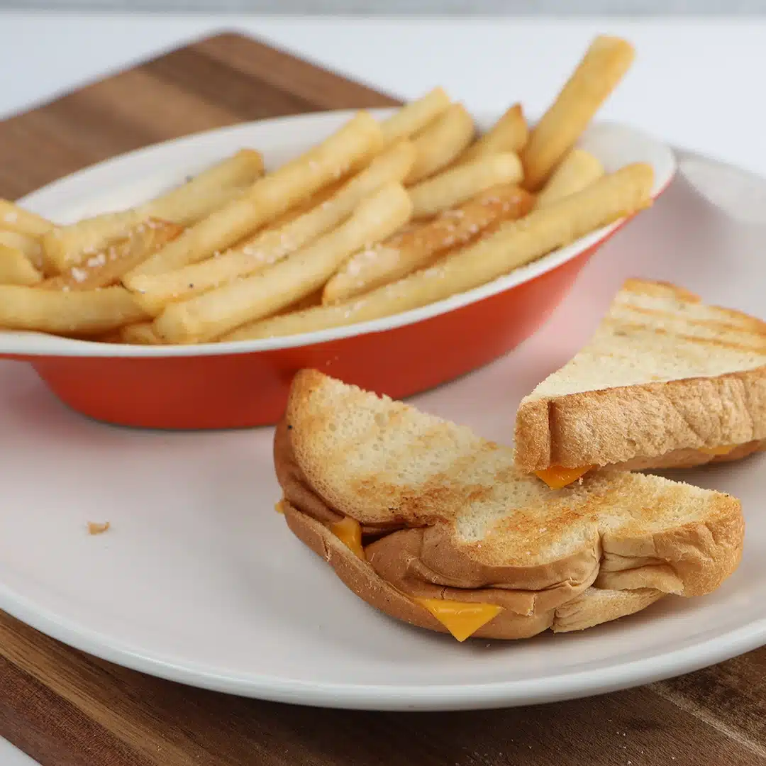 Grilled cheese sandwich cut in half with golden fries in a red dish, on a white plate.