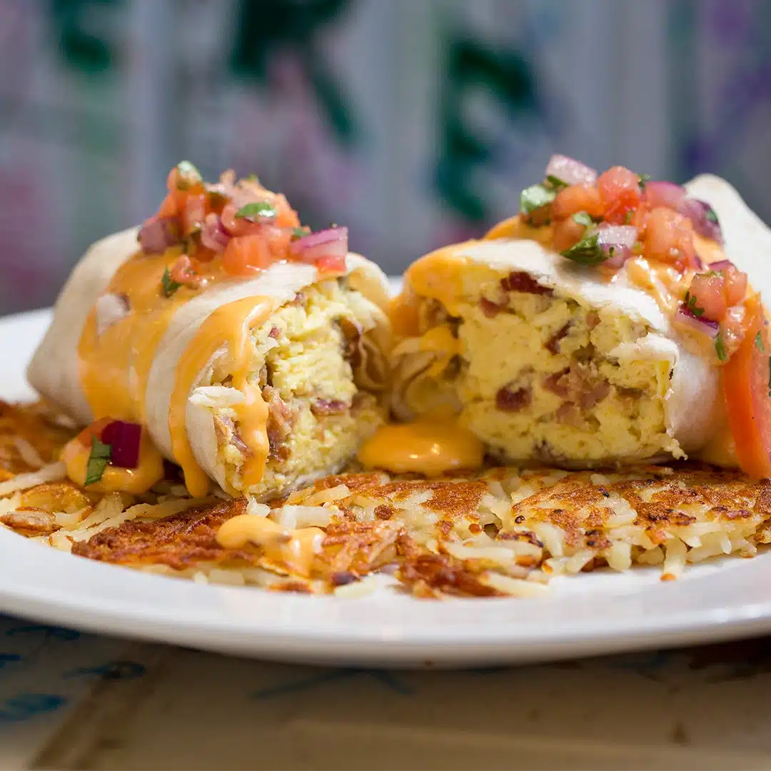 A breakfast burrito split open, revealing eggs and bacon, topped with melted cheese and salsa, served on crispy hashbrowns on a white plate.