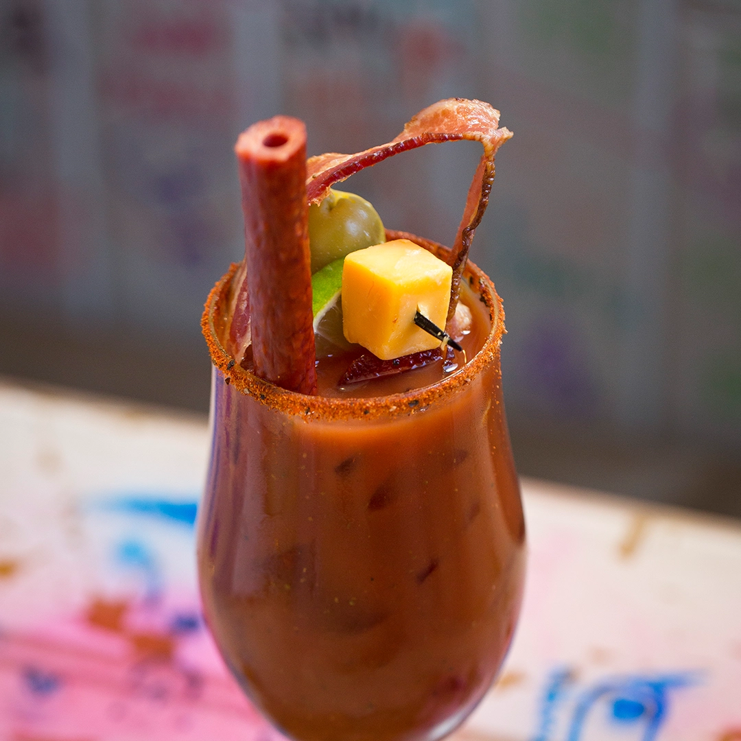 A spicy cocktail garnished with a chili powder rim, skewered cheese, olives, lime, and bacon, creating a bold and festive presentation.