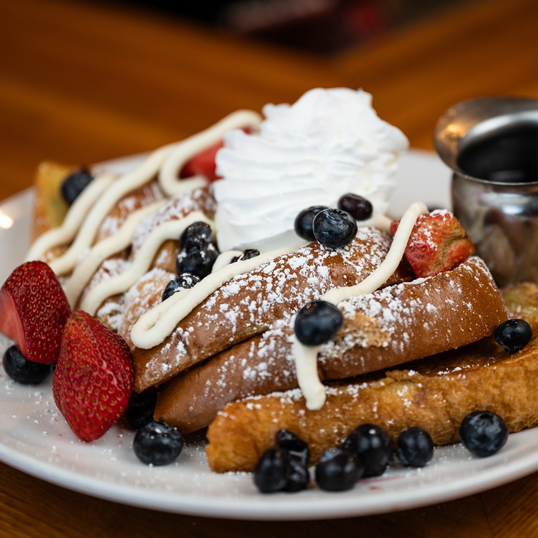 A plate of French toast topped with whipped cream, blueberries, strawberries, powdered sugar, and syrup on the side. 