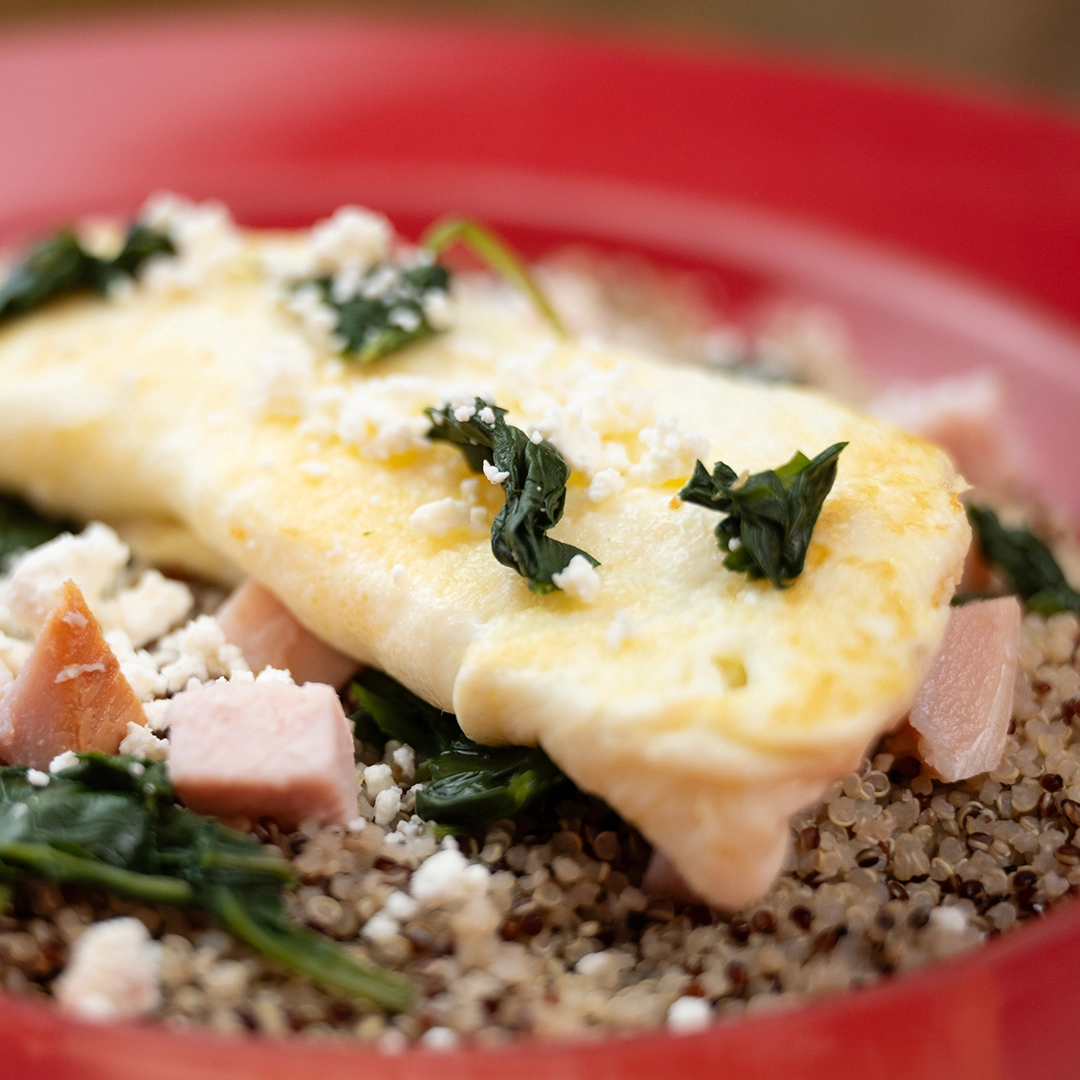 Close-up of a fluffy omelette on a bed of quinoa, garnished with sautéed spinach, diced chicken, and crumbled cheese. Served on a vibrant red plate.
