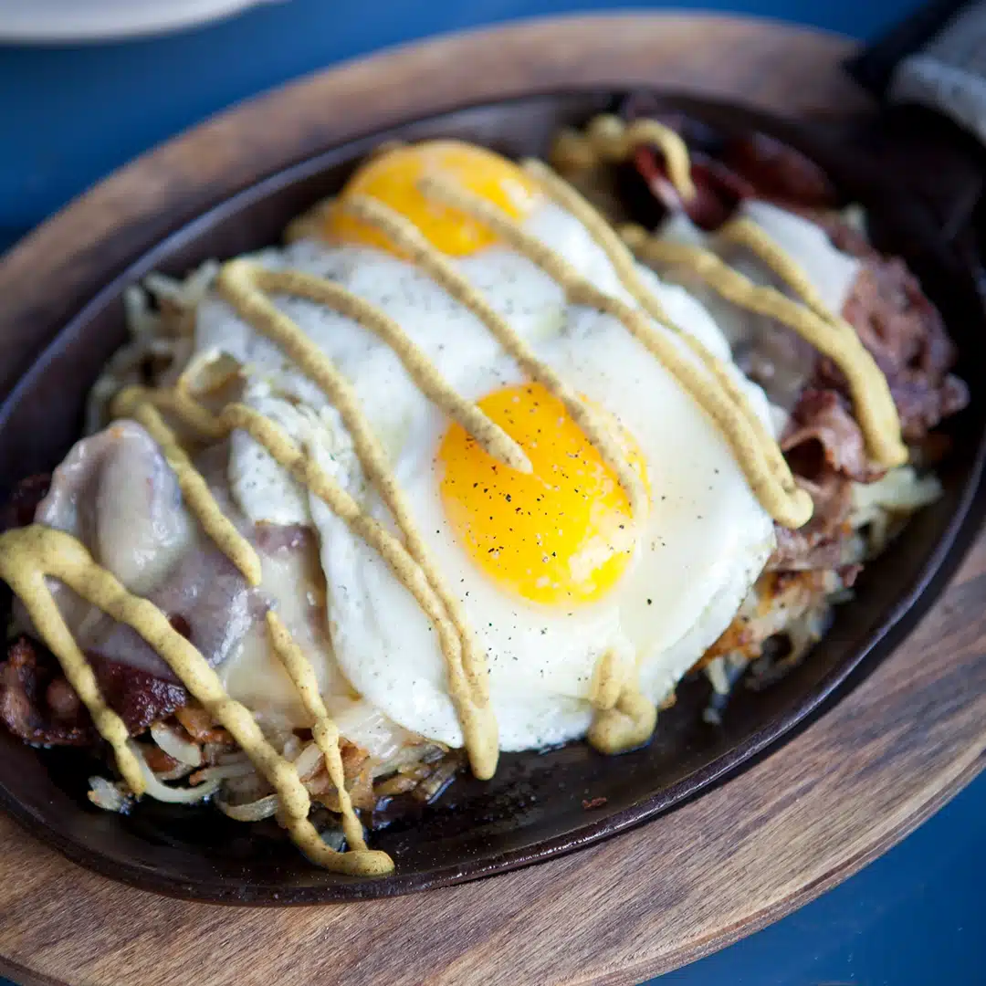 Skillet with melted cheese, crispy hash browns, sliced meat, and two sunny-side-up eggs, drizzled with mustard.