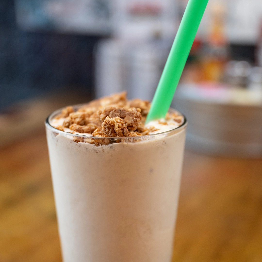 A creamy smoothie topped with granola in a glass, featuring a green straw. The setting has a cozy, inviting atmosphere with blurred background.