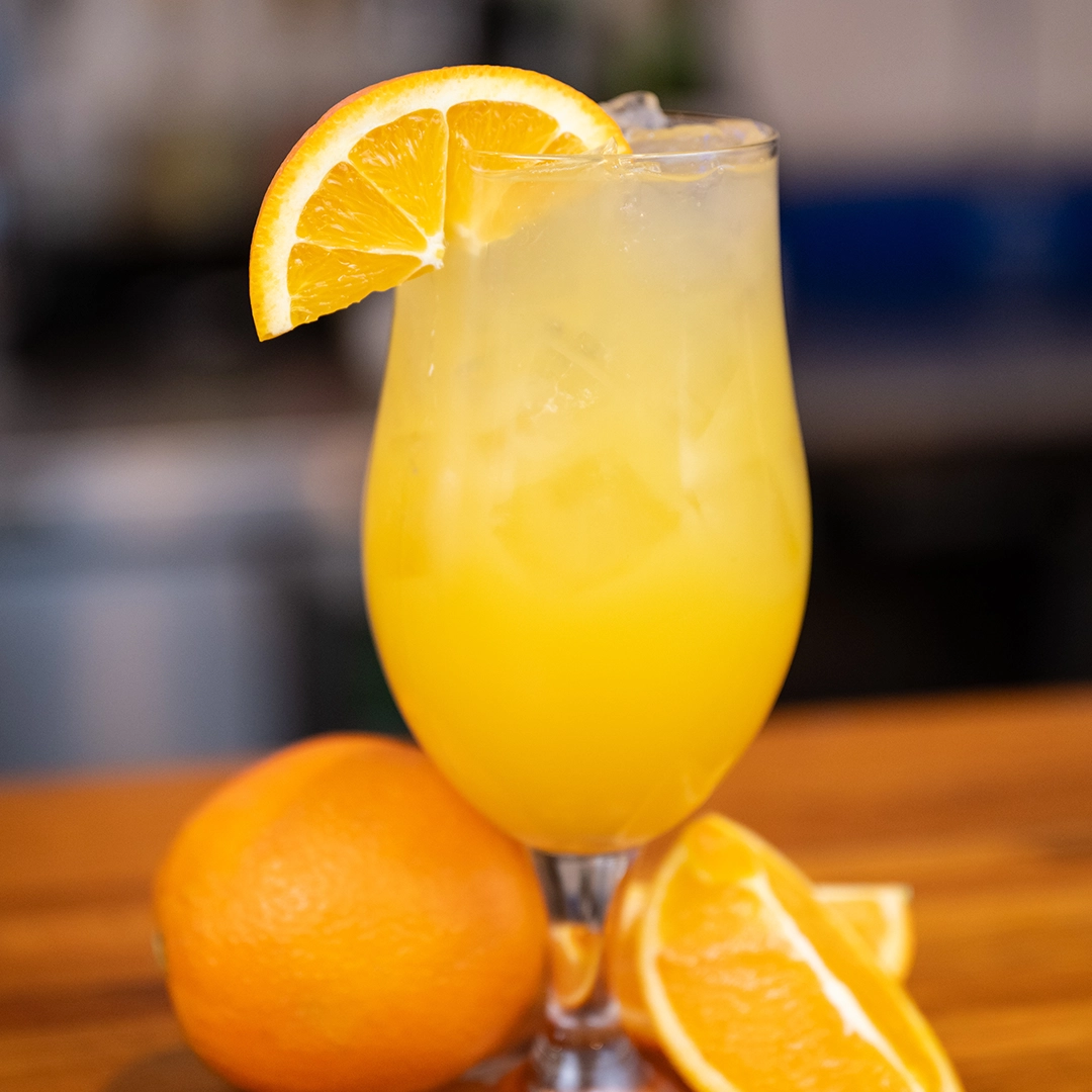A refreshing orange cocktail in a glass with ice, garnished with an orange slice. Whole and sliced oranges sit on a wooden surface beside it.