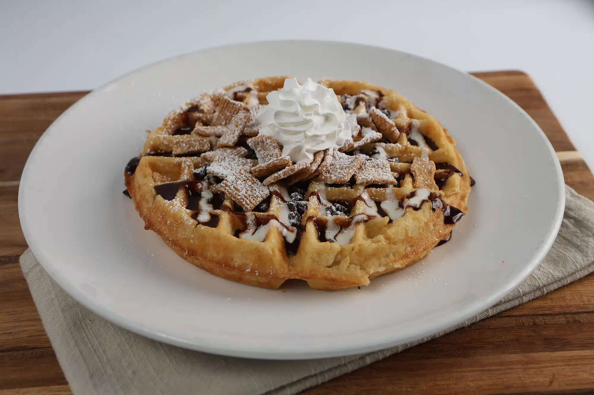 Round waffle topped with whipped cream, cinnamon cereal squares, chocolate drizzle, and powdered sugar.