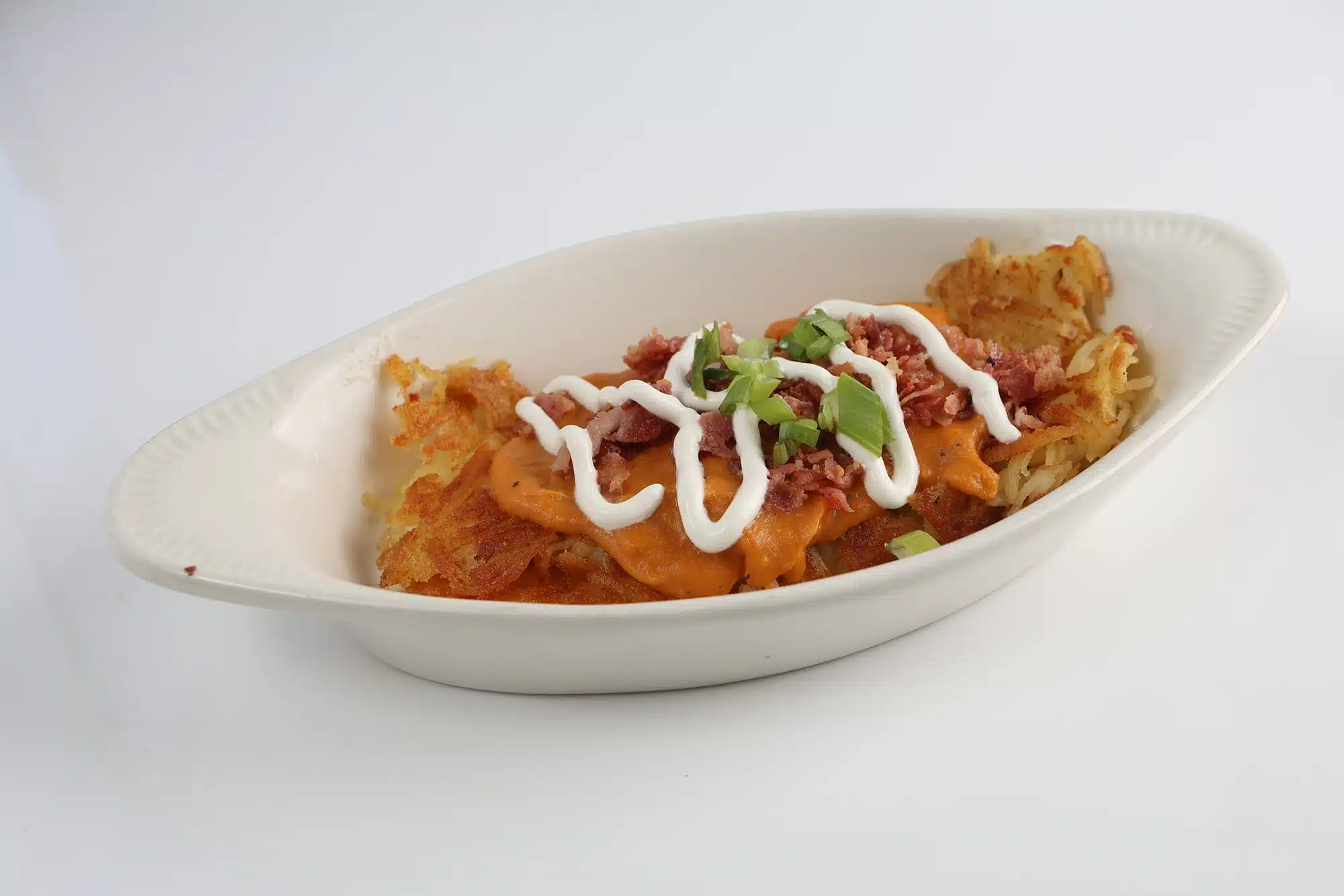 Hash browns topped with spicy orange sauce, sour cream drizzle, crumbled bacon, and chopped green onions in an oval dish.
