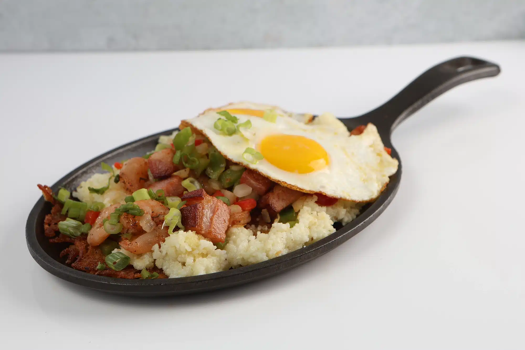 Oval skillet of cheese grits topped with sautéed shrimp, bacon, bell peppers, green onions, and a fried egg.